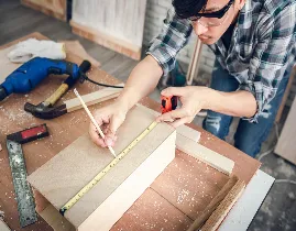 A person in glasses and a plaid shirt measures a piece of wood with a tape measure in a workshop. Tools like a drill, hammer, and saw are visible on the table, and sawdust is scattered around.