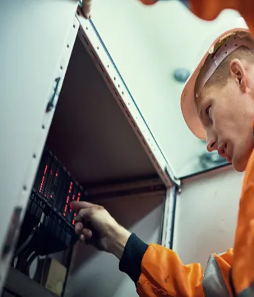 A person wearing an orange safety jacket and helmet is working inside a server room, adjusting equipment on a rack with visible red lights. The room appears to be part of a technical or industrial facility.