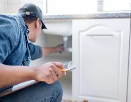 A plumber wearing a cap and denim jacket is working under a kitchen sink. He is holding a wrench and adjusting pipes inside a white cabinet, with tools scattered nearby.