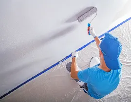 A person wearing a blue shirt and cap, with gloves, paints a wall with a roller. The white wall is partially covered by a large plastic sheet on the floor.