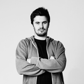 A man with dark hair and a beard stands with arms crossed. He wears a casual jacket over a T-shirt. The background is plain and white, giving the image a simple, monochrome look.