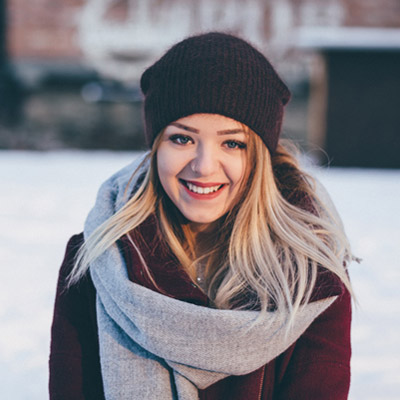 A person with long blonde hair wearing a dark hat and a thick scarf smiles outdoors in a snowy setting. They are dressed warmly in winter clothing, and the background is softly blurred.