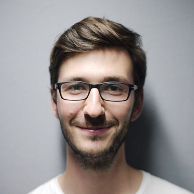 A man with brown hair, glasses, and a short beard smiles while looking at the camera. He is wearing a white shirt and stands in front of a plain gray background.