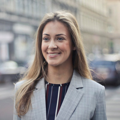 A person with long hair smiles while standing on a city street. They are wearing a plaid blazer over a striped top. The background is slightly blurred, showing buildings and traffic.