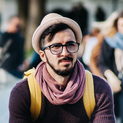 A man wearing a hat, glasses, and a scarf sits outdoors. He has a beard and is wearing a dark sweater and a yellow backpack. Blurred people and trees are in the background.