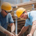 Two construction workers wearing yellow hard hats and blue shirts are focused on a task. They are working with wood at a construction site, with scaffolding and unfinished structures visible in the background.