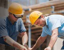 Two construction workers wearing yellow hard hats and blue shirts are focused on a task. They are working with wood at a construction site, with scaffolding and unfinished structures visible in the background.