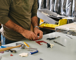 A person in a long-sleeve shirt working at a cluttered table with various tools and materials, including a hammer, pliers, and small pieces of red and white material. Black bags and a yellow container are in the background.