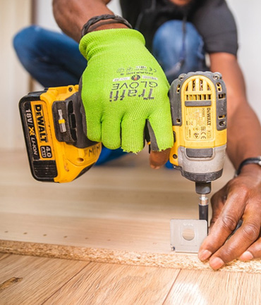 A person wearing bright green gloves operates a yellow power drill while assembling a piece of furniture on a light wooden surface. The focus is on the drill and the hands, suggesting a home improvement or construction project.