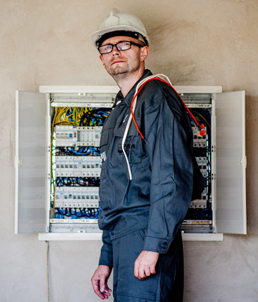 A man in a gray work uniform and white hard hat stands in front of an open electrical panel with various wires and components visible. He has glasses and is looking slightly to the side.