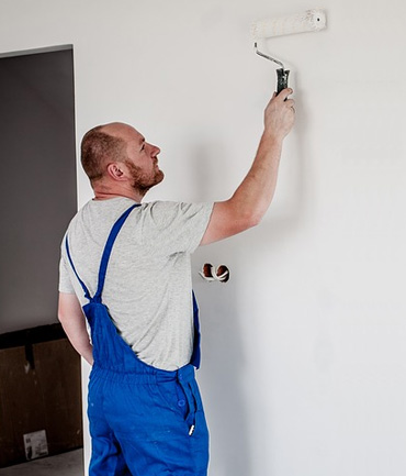 A man in blue overalls is painting a white wall with a roller. He stands in a room with an open doorway, focusing on applying paint evenly. His attention is directed towards the upper section of the wall.