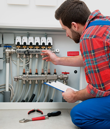 A man in a plaid shirt and blue overalls kneels beside a plumbing manifold, inspecting it while holding a clipboard. Various tools lie on the floor nearby.