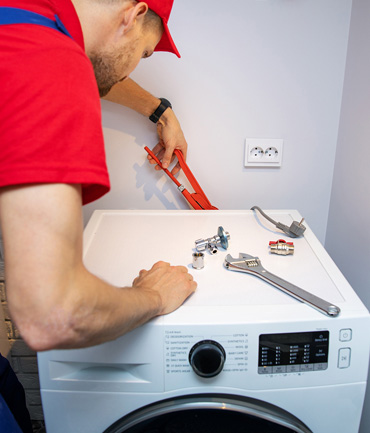 A person in a red cap and shirt installs a washing machine using tools, including a wrench and measuring tape. The machine is near a socket on a light-colored wall.