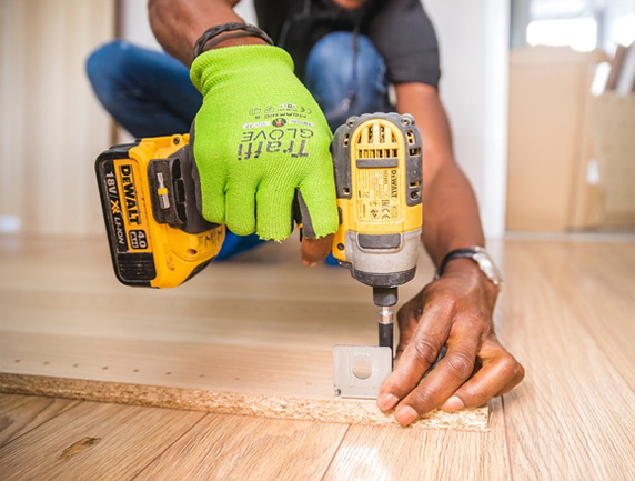 A person wearing bright green gloves uses a DeWalt power drill to screw a metal bracket into a wooden plank. The person is kneeling on a light wood floor, focusing on precise placement of the bracket.