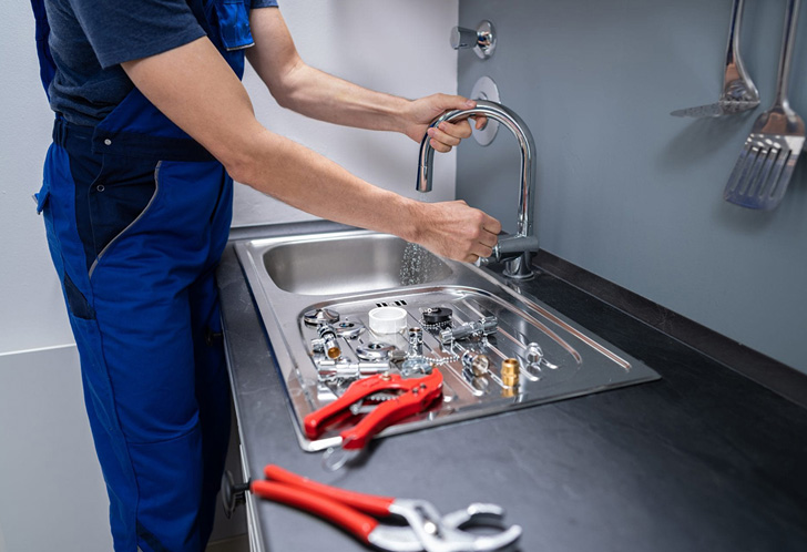 A plumber in blue overalls is working on a kitchen sink. Tools, including pliers and a wrench, are scattered on the countertop. Water is running from the faucet as repairs are underway.