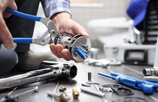 A person using pliers to repair a metal part of a plumbing fixture on a tiled floor, surrounded by various plumbing tools and parts.