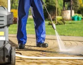 A handyman in blue pants is using a pressure washer to clean a stone patio, the water expertly removing dirt from the surface. Green plants and a garden provide a lush backdrop.