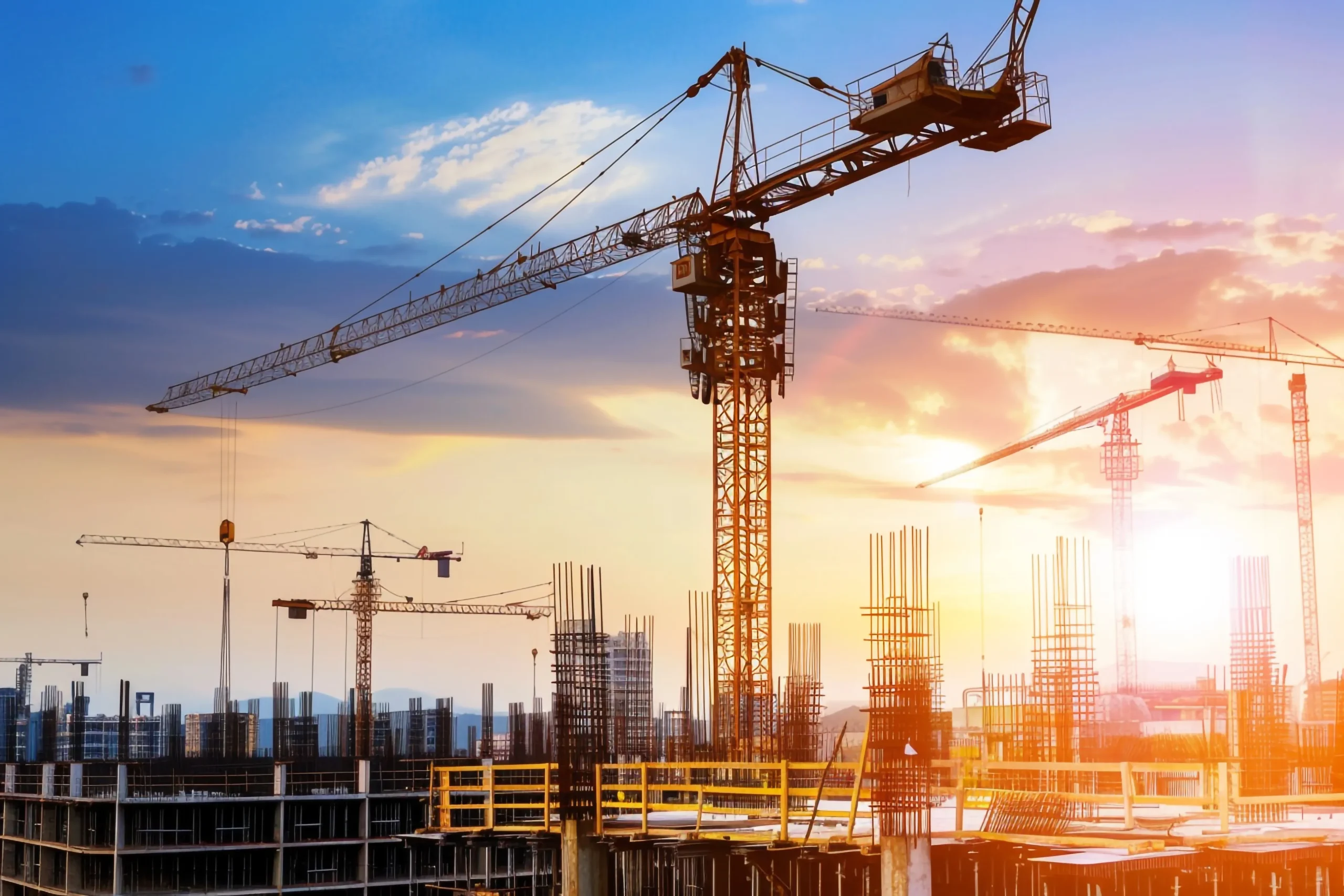 Construction site at sunset, featuring multiple cranes and partially built structures with steel reinforcements. The sky is a blend of orange and blue hues, casting a warm glow over the scene.