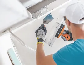 A person wearing a cap and gloves is smoothing plaster on a wall or ceiling with a trowel.