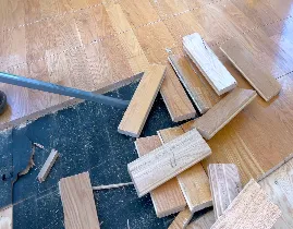 Wooden floorboards are scattered on the ground, some overturned on a black surface. A metal tool, possibly for flooring work, is visible on the left side. The background shows a wooden floor in a room.