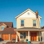 A person stands on a ladder painting the exterior of a two-story house with beige siding. The house has a front porch, and two ladders are set up in the driveway. There are two more houses visible on either side under a clear blue sky.